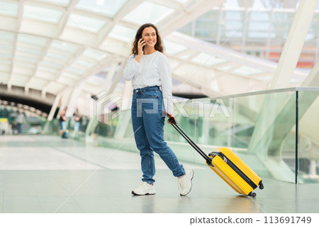 Smiling Woman Talking on Phone While Walking With Suitcase at Airport Terminal 113691749