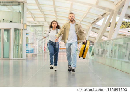 Determined couple sprinting through airport corridor with suitcase, trying to catch flight 113691790