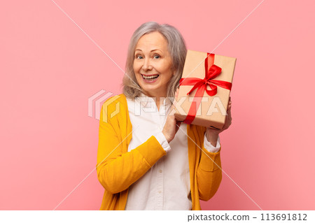 Smiling senior woman holds birthday gift box against pink backdrop 113691812