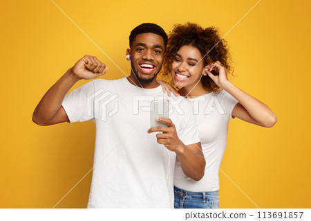 Cheerful black couple sharing headphones with smartphone Cheerful black couple sharing headphones with smartphone 113691857