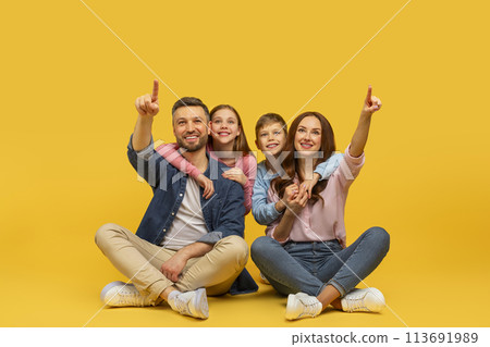 Happy family pointing up sitting together on yellow background Happy family pointing up sitting together on yellow background 113691989