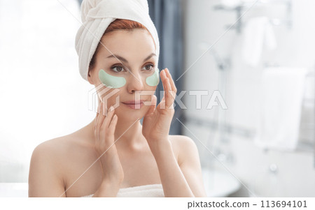 Woman wrapped in towel is examining her face in mirror, focusing on her skin, lips, eyebrows. She smiles at her reflection. Girl with towel on head takes care of her facial skin Woman wrapped in towel is examining her face in mirror, focusing on her skin, lips, eyebrows. She smiles at her reflection. Girl with towel on head takes care of her facial skin 113694101