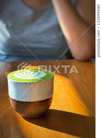 Cup of matcha latte green tea on wooden table with woman in background. Cup of matcha latte green tea on wooden table with woman in background. 113694880