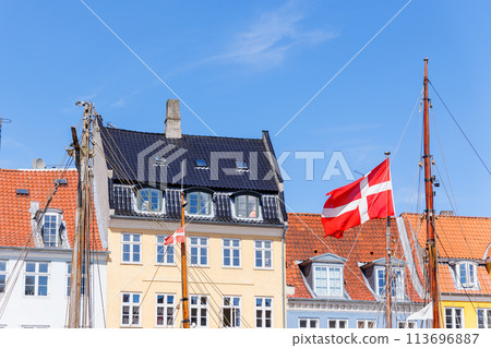 Danish flag waves background Nyhavn colorful house buildings in canal harbor Copenhagen sightseeing tourist travel destination in Denmark on sunny day. Clear blue sky backdrop Danish flag waves background Nyhavn colorful house buildings in canal harbor Copenhagen sightseeing tourist travel destination in Denmark on sunny day. Clear blue sky backdrop 113696887
