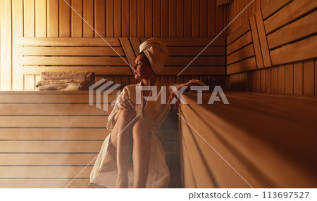 Young woman relaxing and sweating in hot sauna wrapped in towel. Girl In Sauna. Interior of Finnish sauna, classic wooden steam room with hot steam. Russian bathroom. Relax in hot Bathhouse with steam 113697527