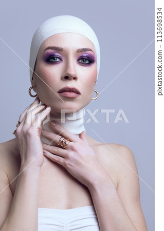Portrait of young beautiful woman in elegant hair accessories, bandage, golden earrings with bright makeup posing on light purple background Portrait of young beautiful woman in elegant hair accessories, bandage, golden earrings with bright makeup posing on light purple background 113698534