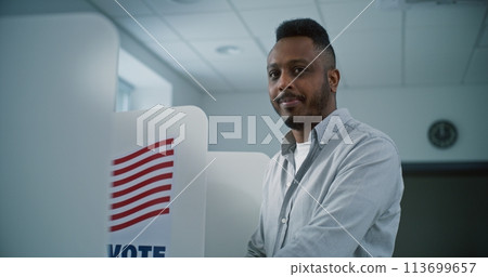 African American man fills out paper ballot in voting booth, than smiles and looks at camera African American man fills out paper ballot in voting booth, than smiles and looks at camera 113699657