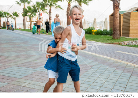Two young boys are laughing and playing a game of wrestling on a paved promenade by the sea 113700173