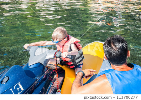 Little boy in a red life jacket seated on a blue jet ski 113700277