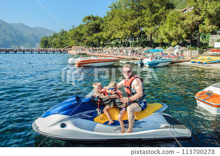 Father with two small sons wearing life jackets is seated on a jet ski near beach Father with two small sons wearing life jackets is seated on a jet ski near beach 113700278