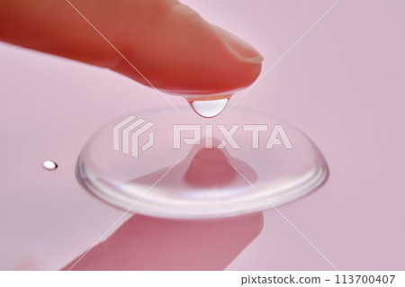 A finger touches a drop of cosmetic product on a pink background. 113700407