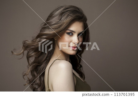 Elegant woman with long healthy wavy hair, cute smile and make-up looking at camera on brown studio Elegant woman with long healthy wavy hair, cute smile and make-up looking at camera on brown studio 113701395