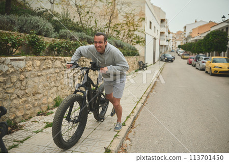European young man pushing his electric mountain bike up the street. Active lifestyle. Sport. Adventure travel on bicycle. Using el-bicycle ad ecologically friendly mode of urban transport 113701450