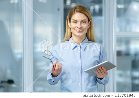 Beautiful businesswoman smiling at camera with stylish eyeglasses and electronic tablet in hands. Middle age confident entrepreneur getting ready for new working day in stylish corporate building. Beautiful businesswoman smiling at camera with stylish eyeglasses and electronic tablet in hands. Middle age confident entrepreneur getting ready for new working day in stylish corporate building. 113701945
