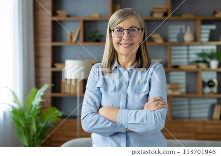Confident female pensioner in pastel blouse standing with arms crossed on chest and smiling at camera. Mature lady in spectacles posing at stylish apartment interior and enjoying holiday indoors. Confident female pensioner in pastel blouse standing with arms crossed on chest and smiling at camera. Mature lady in spectacles posing at stylish apartment interior and enjoying holiday indoors. 113701946