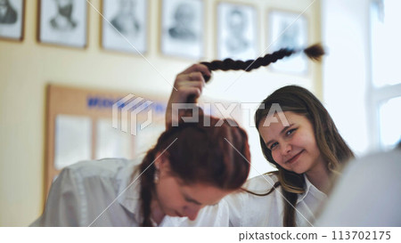 Fun schoolgirls in the classroom. Girl playing with her friend's pigtail. Fun schoolgirls in the classroom. Girl playing with her friend's pigtail. 113702175