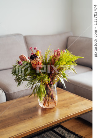 Vase with exotic protea flowers bouquet on coffee table with blurred background of modern cozy light living room with gray couch sofa. Open space home interior design. Elegant home decor. Copy space. Vase with exotic protea flowers bouquet on coffee table with blurred background of modern cozy light living room with gray couch sofa. Open space home interior design. Elegant home decor. Copy space. 113702474