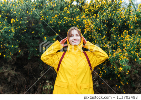 Smiling young woman in a yellow raincoat and hood enjoying walk on background of blooming bush with flowers. Travel concept. Hiking in any weather. Girl on a journey. Relaxing, personal fulfillment Smiling young woman in a yellow raincoat and hood enjoying walk on background of blooming bush with flowers. Travel concept. Hiking in any weather. Girl on a journey. Relaxing, personal fulfillment 113702488