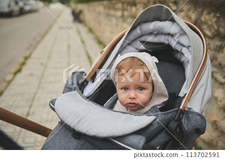 Portrait of an adorable European baby boy with hood on his head, looking at the camera, sitting in a baby pram outdoors Portrait of an adorable European baby boy with hood on his head, looking at the camera, sitting in a baby pram outdoors 113702591