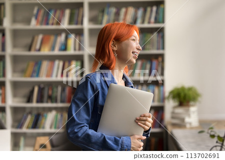 Happy red-headed girl holding laptop standing in college library 113702691