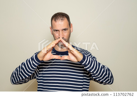 Hispanic man with a beard with his fingertips together in an expression of evil, cunning and cold planning a crime. Isolated on beige background. 113702762