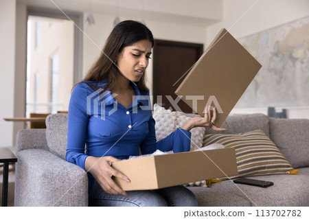 Upset concerned young Indian customer woman opening logistic cardboard box 113702782
