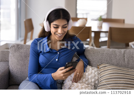 Cheerful young Indian girl in big white headphones using smartphone 113702918