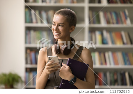 Smiling pensive student girl standing in library with smartphone 113702925