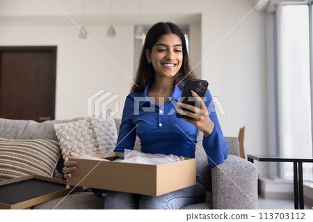 Cheerful Indian girl typing on smartphone over open box Cheerful Indian girl typing on smartphone over open box 113703112