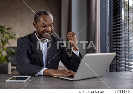 A man in a suit is sitting at a desk with a laptop and a tablet. He is smiling and he is happy 113703420