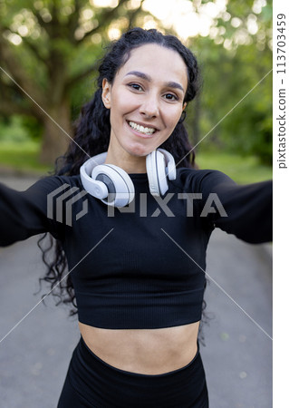 A smiling Hispanic woman enjoys a workout break in the park, taking a selfie with her headphones around her neck. She wears a black sports outfit. 113703459