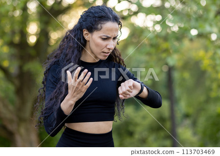 A confused Hispanic woman in workout attire checks her watch during an exercise session in a park, expressing uncertainty and slight frustration. A confused Hispanic woman in workout attire checks her watch during an exercise session in a park, expressing uncertainty and slight frustration. 113703469