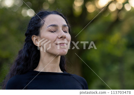 A serene Hispanic woman closes her eyes, enjoying a tranquil moment outdoors surrounded by greenery, feeling calm and peaceful. 113703478