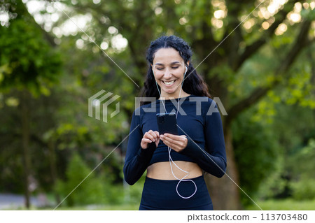 A joyful Hispanic woman listens to music on her smartphone while running in a lush green park. She wears a black sports outfit and exudes happiness and health. 113703480