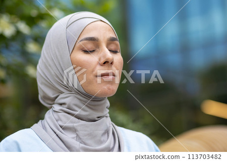 A serene portrait of a Muslim woman meditating with closed eyes, wearing a hijab, in a verdant urban park, exuding a sense of peace and contentment. 113703482
