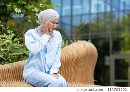 A young woman wearing a hijab experiences discomfort due to ear pain. She is sitting on a wooden bench, expressing concern, with a modern building in the background. A young woman wearing a hijab experiences discomfort due to ear pain. She is sitting on a wooden bench, expressing concern, with a modern building in the background. 113703502