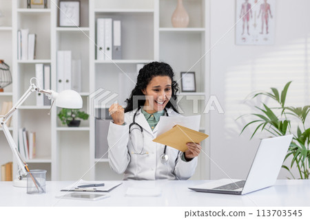 A cheerful mature female doctor excitedly celebrates while reading good news from an envelope in her bright, organized clinic office, exuding professionalism and happiness. 113703545
