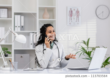 A cheerful female doctor with curly hair happily talking on the phone while looking at her laptop in a well-lit, organized office. 113703564