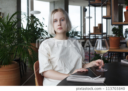 Beautiful young woman sitting at table holding in hand glass of drink at restaurant. Blonde pretty lady 113703840