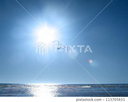 kites flies at the background of blue sea and sky, and bright sun with lens flare, and glowing light, kitesurfing  (the sail pulls along the waves of a man on the board) 113705631