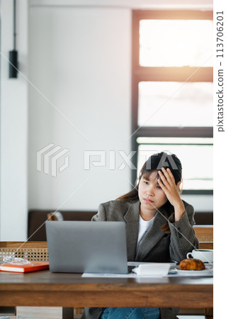 Woman seated at a cafe table, expressing concern while looking at her laptop screen, with hand on forehead. Woman seated at a cafe table, expressing concern while looking at her laptop screen, with hand on forehead. 113706201