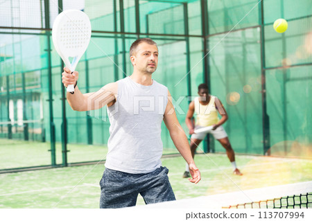 Male players playing padel in a padel court outdoor behind net 113707994