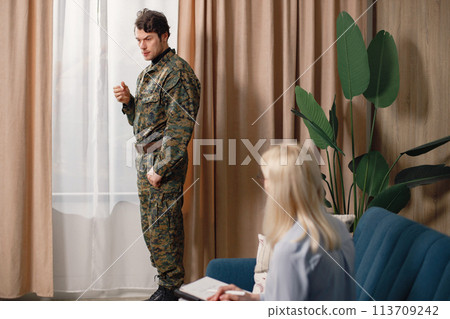 Man in military uniform standing near windows, woman psychiatrist sitting on a couch. Soldier and psychiatrist talking during therapy session. Male warrior with ptds treating his mental disorder. Man in military uniform standing near windows, woman psychiatrist sitting on a couch. Soldier and psychiatrist talking during therapy session. Male warrior with ptds treating his mental disorder. 113709242