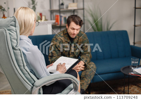 Soldier and psychiatrist sitting on couch during therapy session. Man wearing military uniform. Male warrior with ptds talking to psychiatrist. 113709248