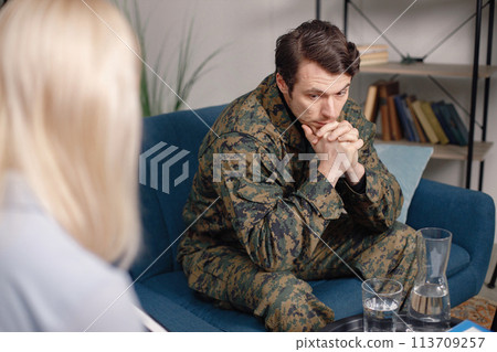 Cropped photo of a soldier and psychiatrist sitting on couch during therapy session. Man wearing military uniform. Male warrior with ptds talking to psychiatrist. 113709257