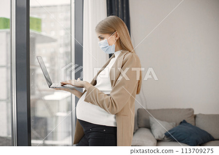 Portrait of pregnant woman typing on a laptop while standing near windows in modern office. Pregnant businesswoman using a laptop in her office. Blonde woman wearing beige jacket and medical face mask 113709275