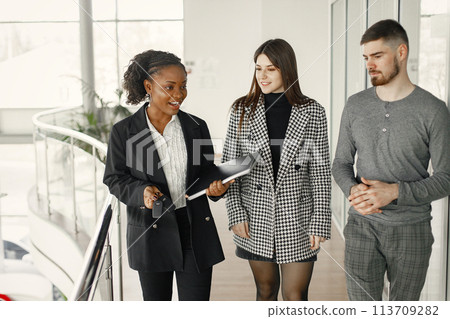 Professional car dealer standing at auto showroom and holding clipboard and car key. Man and woman want to buy a car. Black woman wearing a black costume and white shirt. Professional car dealer standing at auto showroom and holding clipboard and car key. Man and woman want to buy a car. Black woman wearing a black costume and white shirt. 113709282