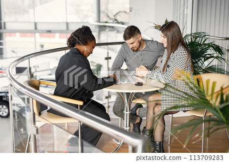 Professional car dealer sitting at the table with clipboard and documents. Two customers behind looking on a documents. Caucasian man and woman in auto showroom. Professional car dealer sitting at the table with clipboard and documents. Two customers behind looking on a documents. Caucasian man and woman in auto showroom. 113709283