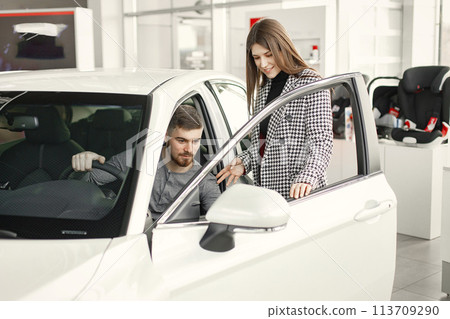 Beautiful young couple standing at the dealership choosing the car to buy. Man sitting inside a car. Couple wearing grey clothes. 113709290