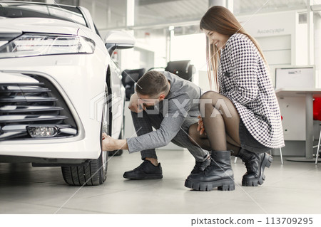 Beautiful young couple standing at the dealership choosing the car to buy. Man and woman looking on a tires. Couple wearing grey clothes. Beautiful young couple standing at the dealership choosing the car to buy. Man and woman looking on a tires. Couple wearing grey clothes. 113709295